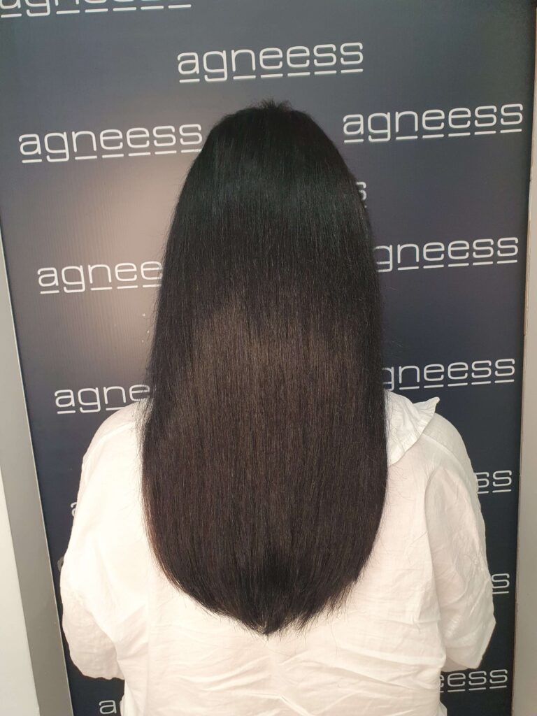 Widok od tyłu na głowę kobiety po zabiegu przedłużania włosów w salonie Agneess. Włosy są gładkie, zdrowo wyglądające, o ciemnobrązowym kolorze, sięgające poniżej ramion. Tło stanowi ściana z białym logo salonu Agneess na ciemnym tle