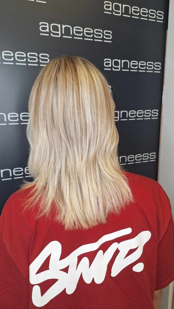 Widok z tyłu na włosy klientki po koloryzacji i strzyżeniu w salonie fryzjerskim Agneess. Włosy są świeżo obcięte i mają wykonany balayage w odcieniach blond, z naturalnymi przejściami kolorystycznymi, które dodają fryzurze objętości i blasku. Klientka ubrana w czerwoną koszulkę z białym napisem, tło z logo Agneess.