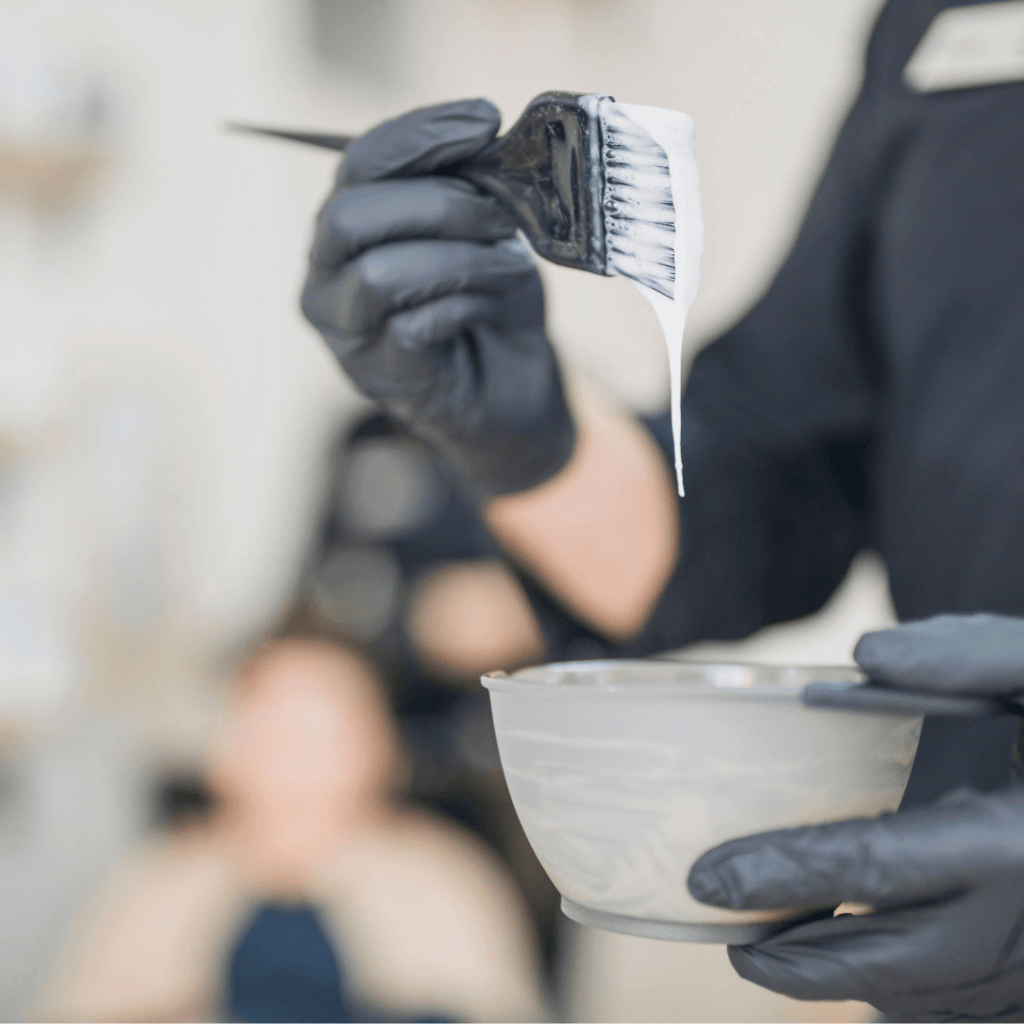 ręka mężczyzny ubrana w czarną lateksową rękawiczkę trzyma miskę z farbą fryzjerską oraz pędzelek do farbowania włosów